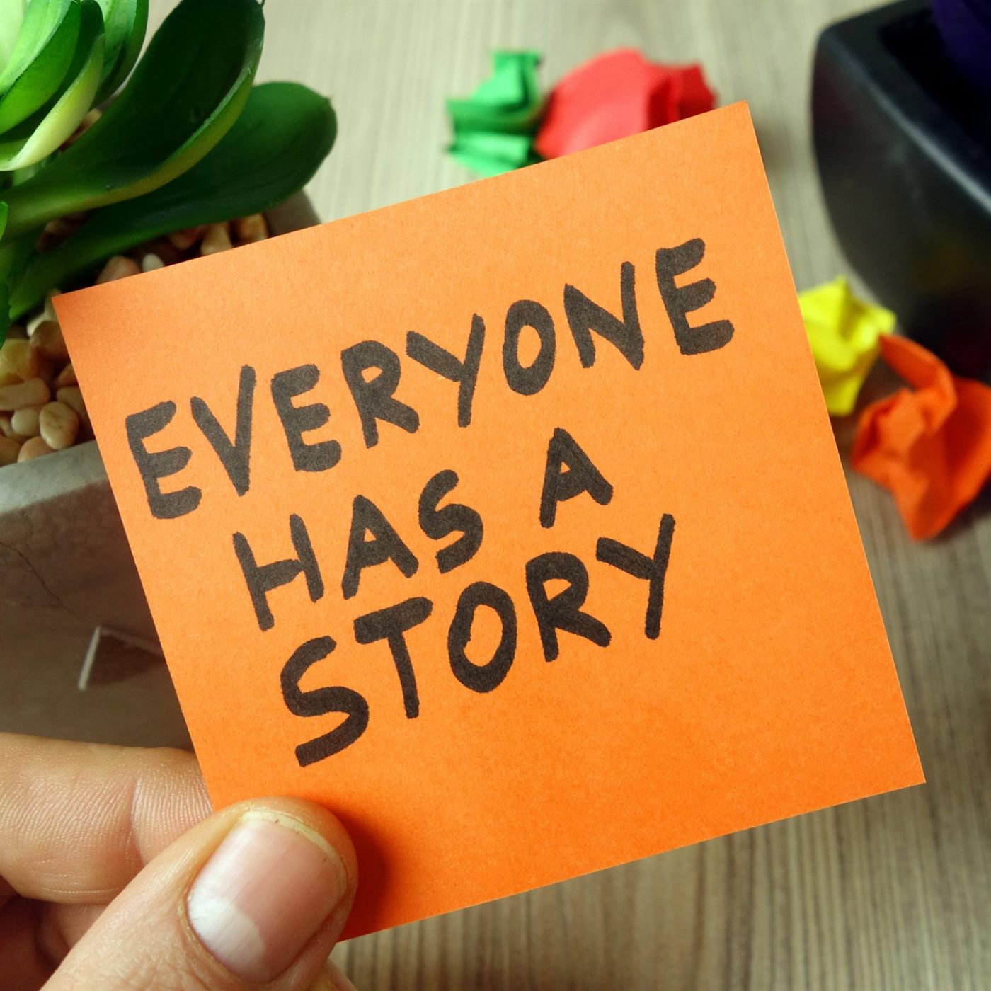 An orange sticky note reading “Everyone has a story”, held in a hand
